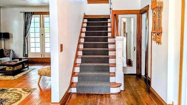 Un escalier recouvert d’une moquette sombre, avec des marches en bois et des moulures blanches. Un lustre est suspendu au-dessus de l’escalier, et une porte ornée d’un cadre décoratif se trouve sur la droite.