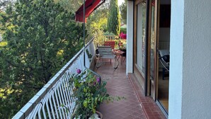 A balcony with a red awning, potted plants, a metal railing, and a view of trees and a garden.