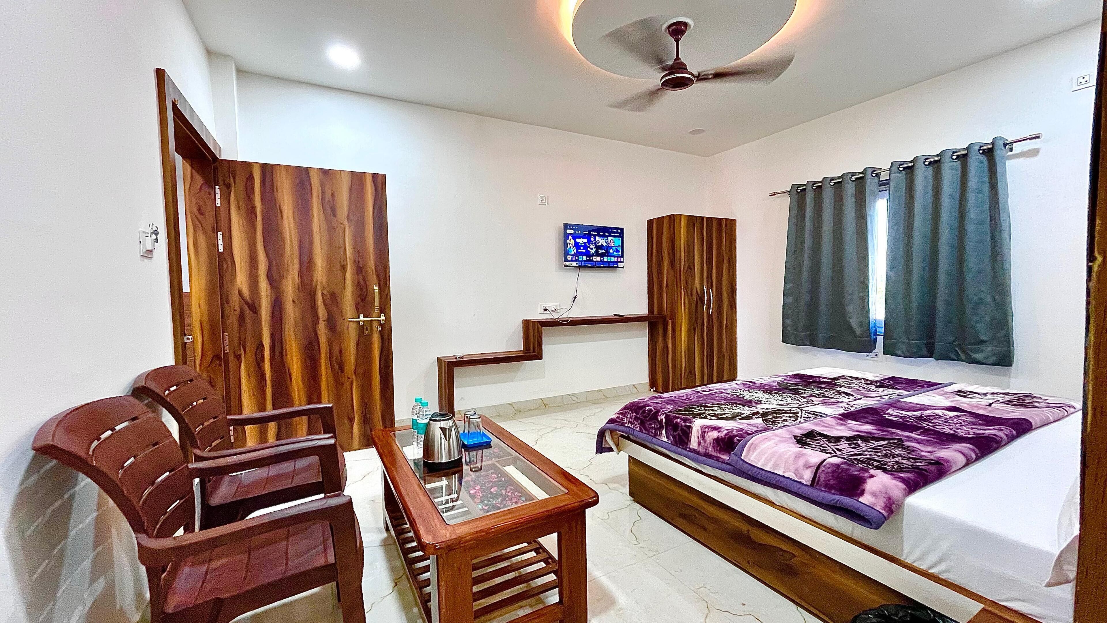A bedroom with a bed, wooden wardrobe, a desk with a chair, a television on the wall, and a ceiling fan.