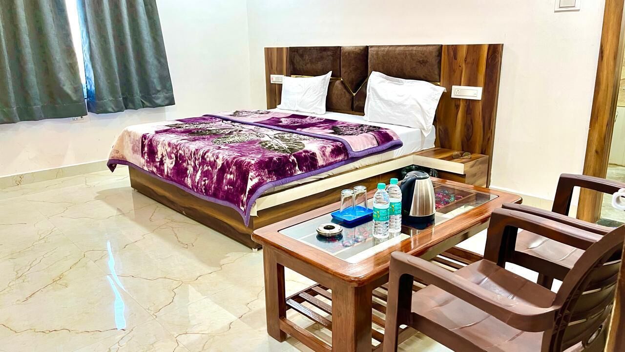 Quarto de hotel com cama, mesa de cabeceira de madeira, mesa e cadeiras, e janela com cortinas.