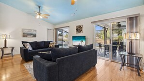 A spacious living area with a dark gray sofa, a coffee table, and a ceiling fan. There is a large window providing a view of the outdoors.