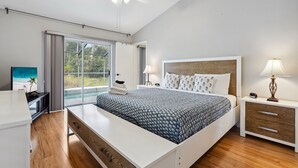 A modern bedroom with a large bed, a desk, and a nightstand. There is a window with a view of greenery outside.