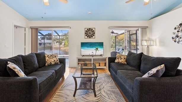 A living room with two dark sofas, a glass coffee table, a flat-screen TV, ceiling fans, and large windows.