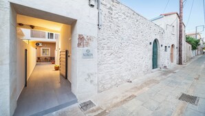 A narrow street with a building labeled "NIKOS" featuring a covered entrance and a small seating area with chairs and potted plants. - Nikos StoneHouse family home, located in the center of Prines (Rethymno)