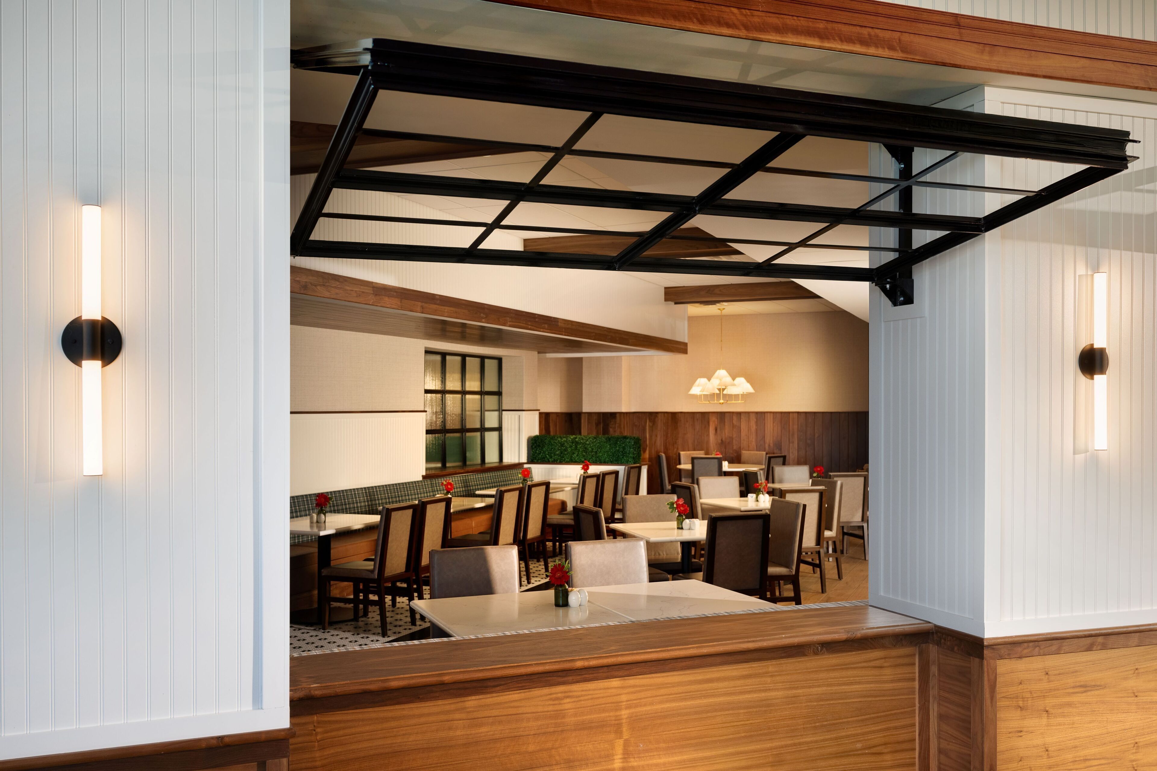 Uma zona de refeições de hotel moderna com mesas e cadeiras de madeira, teto alto com vigas aparentes e iluminação embutida na parede.