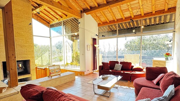 A spacious living room with a high wooden ceiling, a fireplace, large windows, and a red sofa set. - Le SIG - Swimming pool - 6 rooms (Piolenc)