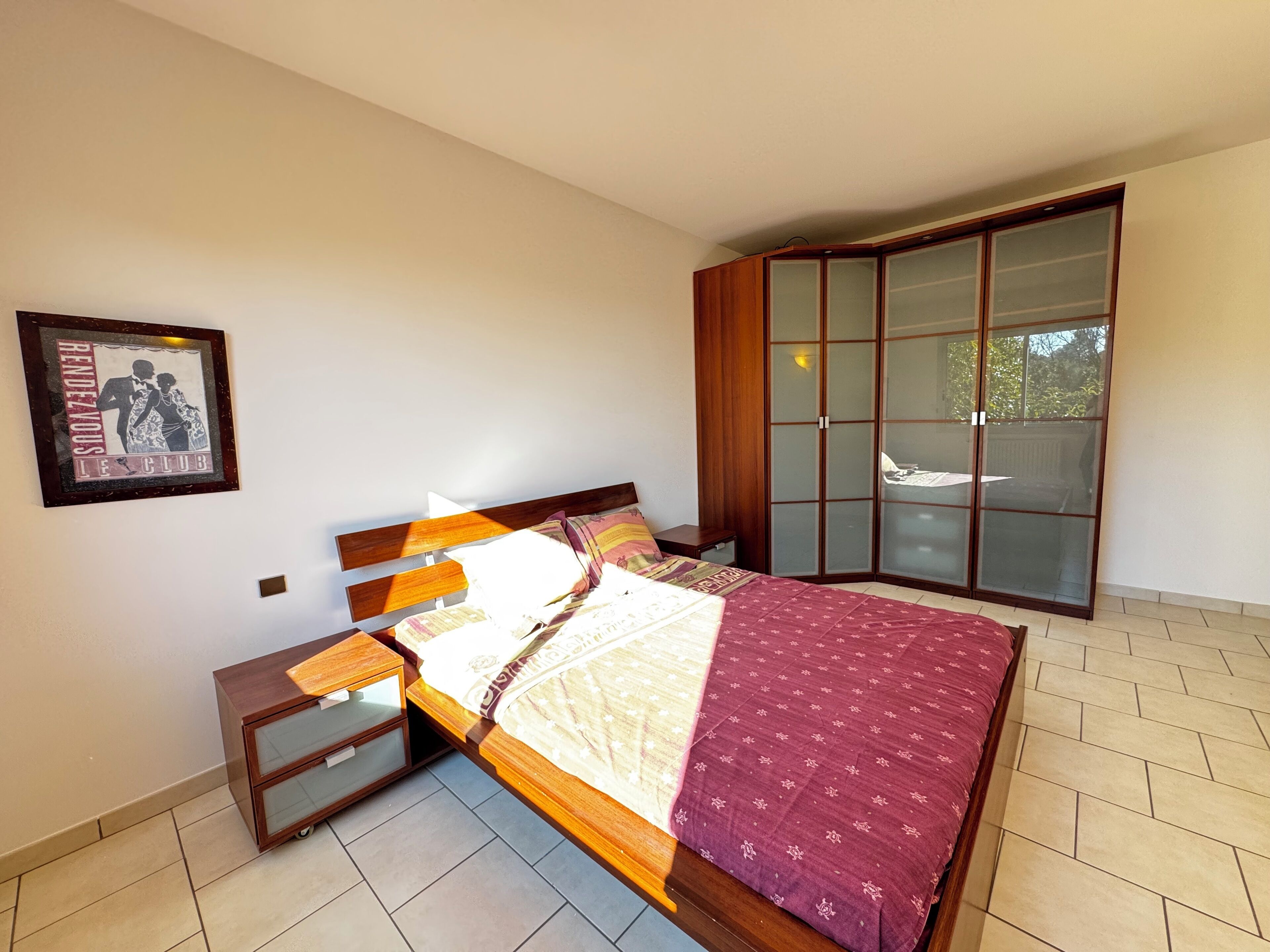 Een slaapkamer met een bed, houten nachtkastjes, een garderobe met glazen deuren en een lijst met een afbeelding aan de muur.