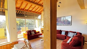 A spacious living area with wooden beams, a red sofa, a blue sofa, and a large window providing a view of the outdoors. - Le SIG - Swimming pool - 6 rooms (Piolenc)
