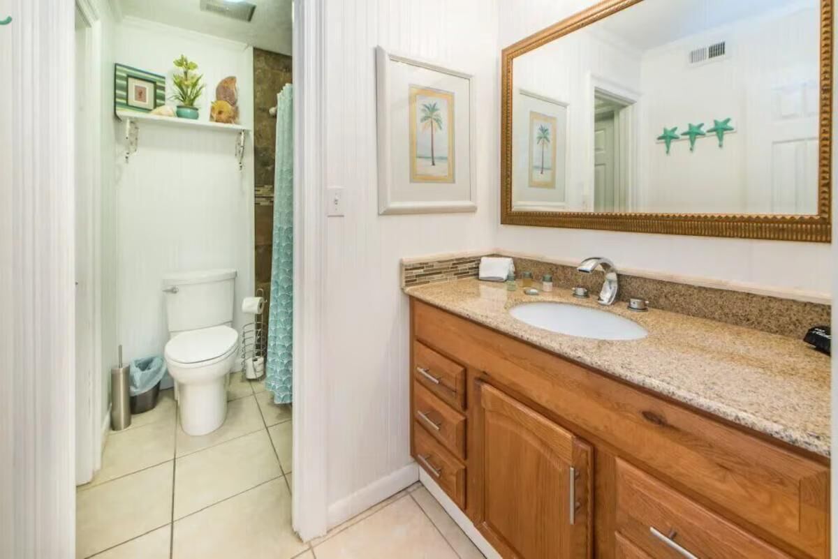 A bathroom with a toilet, sink, and mirror. There are framed pictures on the wall and a shelf with decorative items.
