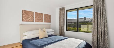 Ein Schlafzimmer mit einem Bett, einer Holzbank, gemusterten Vorhängen und einem Fenster mit Blick auf Grünfläche.