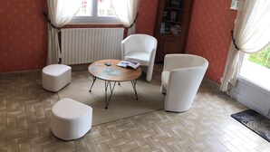 A room with a herringbone floor, a round wooden table with cards and a book, two white armchairs, a white ottoman, a radiator, and a wooden cabinet with books. - Family home between sea and countryside for 6 people (aubigny)