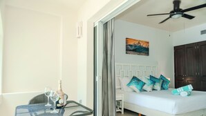 Ein Schlafzimmer mit einem Bett, einem Nachttisch, einem Sessel, einem Tisch mit Flasche und Gläsern sowie einem Deckenventilator.