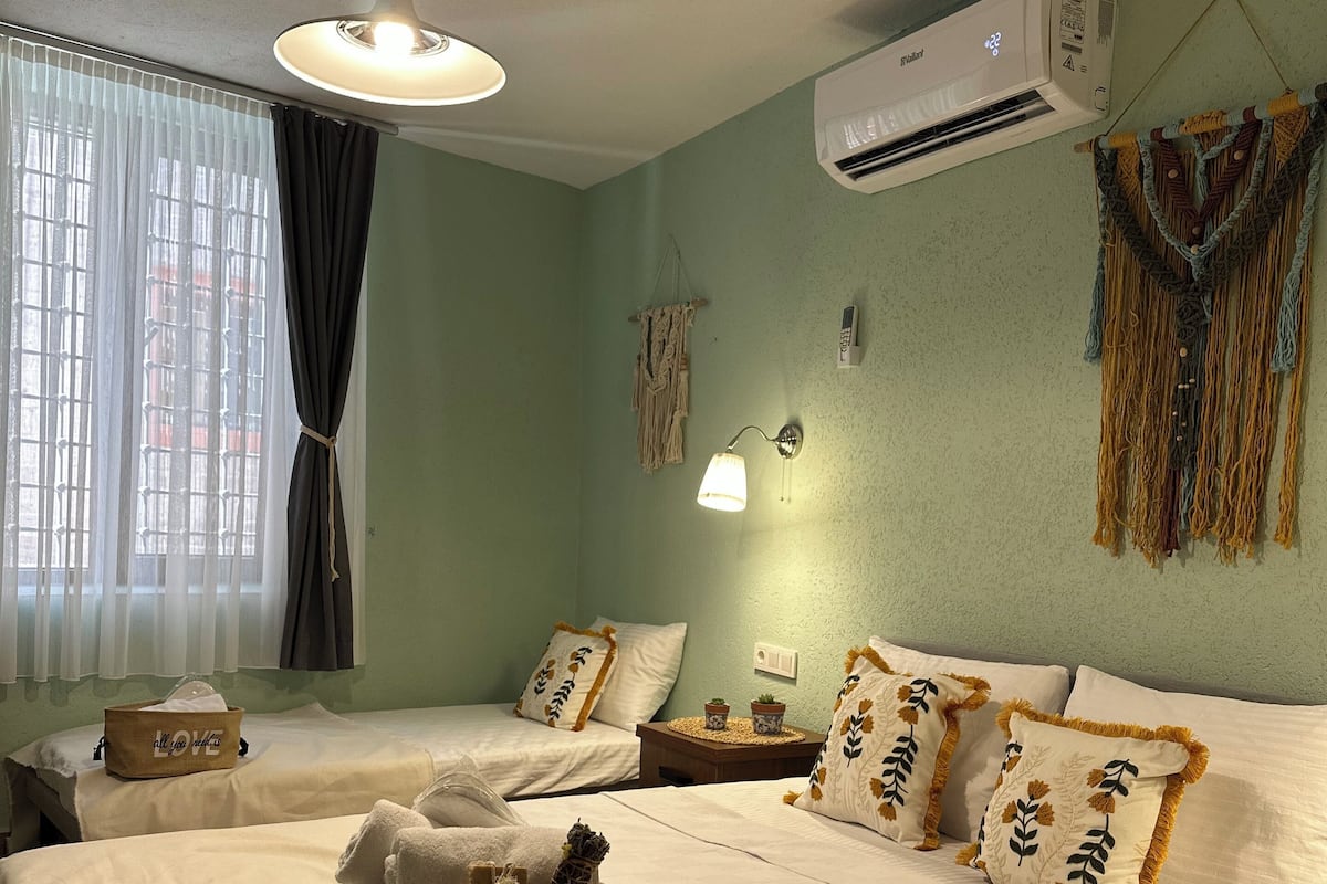 A hotel room with two beds, a ceiling fan, an air conditioner, a bedside lamp, a wall-mounted phone, and a decorative macrame piece.