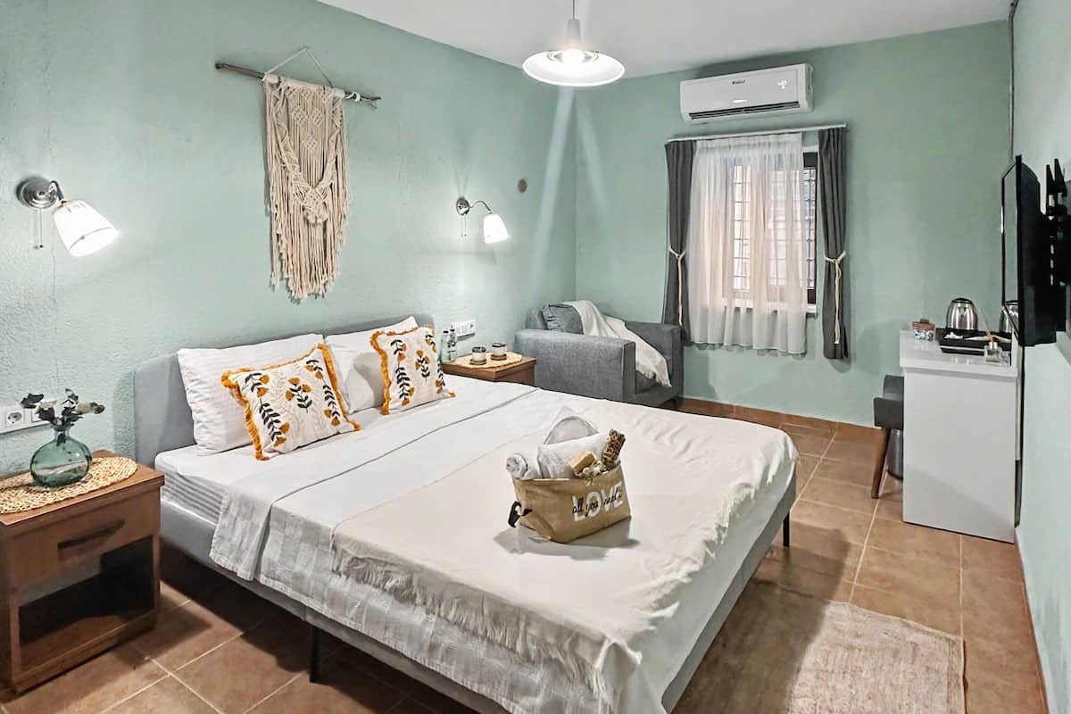 A bedroom with a bed, bedside tables, a desk, a chair, a hanging macrame wall decor, a ceiling fan, and an air conditioning unit.