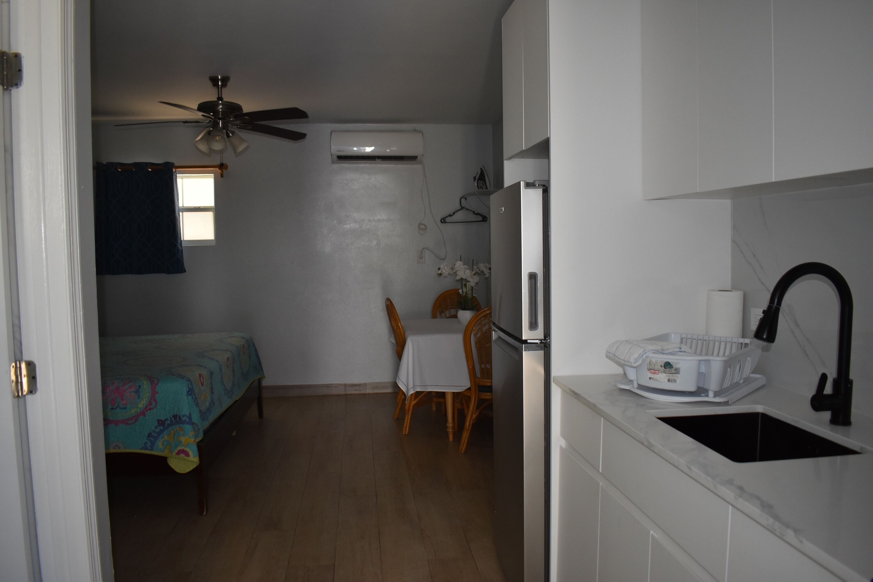 Una habitación compacta con cama, una pequeña mesa de comedor, ventilador de techo, aire acondicionado, refrigerador blanco y pileta de cocina.