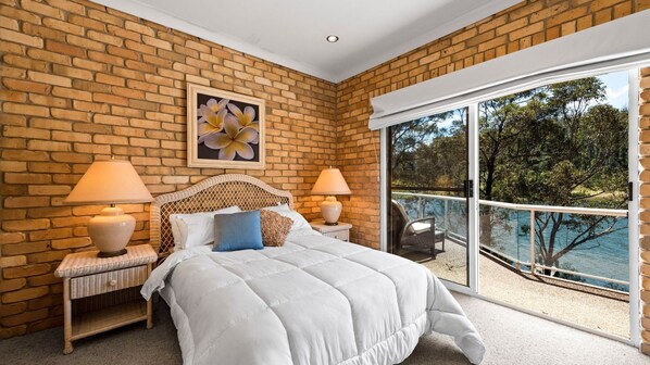 A bedroom with a brick wall, a large bed, two bedside lamps, a framed picture, and a balcony with a view of trees and water.
