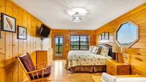 A wooden cabin bedroom with a bed, a rocking chair, a wooden nightstand, and a large window. - 3 miles to Dollywood! Mountain views! Hot tub! Family fun! (Sevierville)