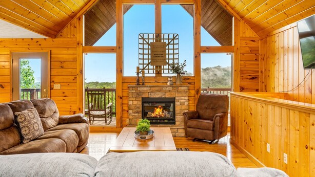 A cozy log cabin interior with a fireplace, wooden furniture, and a large window offering a scenic view. - 3 miles to Dollywood! Mountain views! Hot tub! Family fun! (Sevierville)