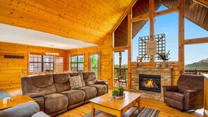 A cozy log cabin interior with a fireplace, wooden furniture, and large windows offering views of the outdoors. - 3 miles to Dollywood! Mountain views! Hot tub! Family fun! (Sevierville)