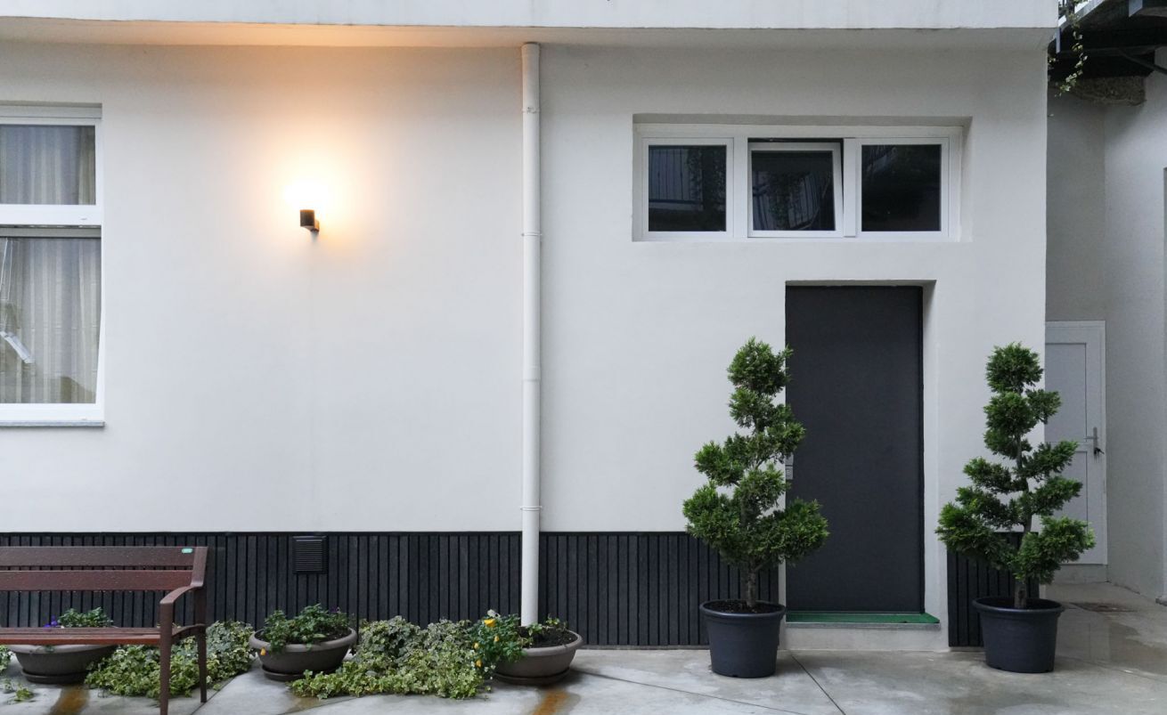 A modern building with a white facade, a dark door, and three windows. There are potted plants and a bench outside.