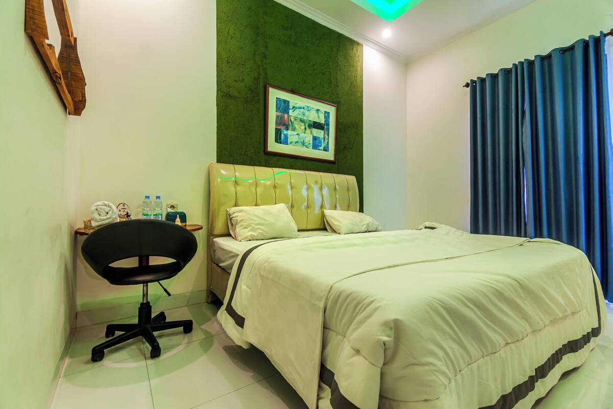 A hotel room with a bed, a desk with a chair, a green accent wall, and a framed picture above the bed.