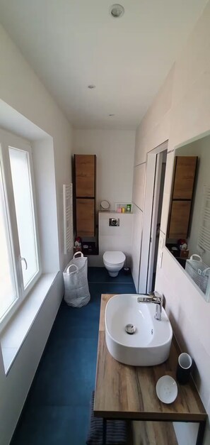 A narrow bathroom with a white sink, a toilet, and a window. - Under the eye of the bell tower (peymeinade)