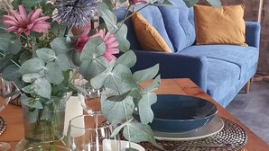 A rustic stone wall, a blue sofa with a mustard cushion, and a wooden table with a vase of flowers and a plate. - Under the eye of the bell tower (peymeinade)
