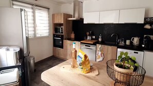 A modern kitchen with a stainless steel refrigerator, a built-in oven, a microwave, and a dishwasher. There is a wooden countertop with a cutting board, a bottle of olive oil, and a bag of flour. A potted plant is on the countertop. - Under the eye of the bell tower (peymeinade)