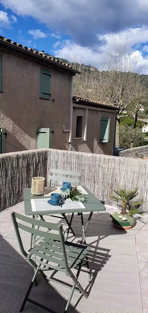 A patio with a table and chairs, a potted plant, and a building in the background. - Under the eye of the bell tower (peymeinade)