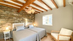 Un dormitorio con dos camas, una pared de piedra, vigas de madera, un sillón mecedora y una ventana.