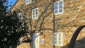 Un edificio de piedra con varias ventanas y una puerta, rodeado de vegetación y un área pavimentada.
