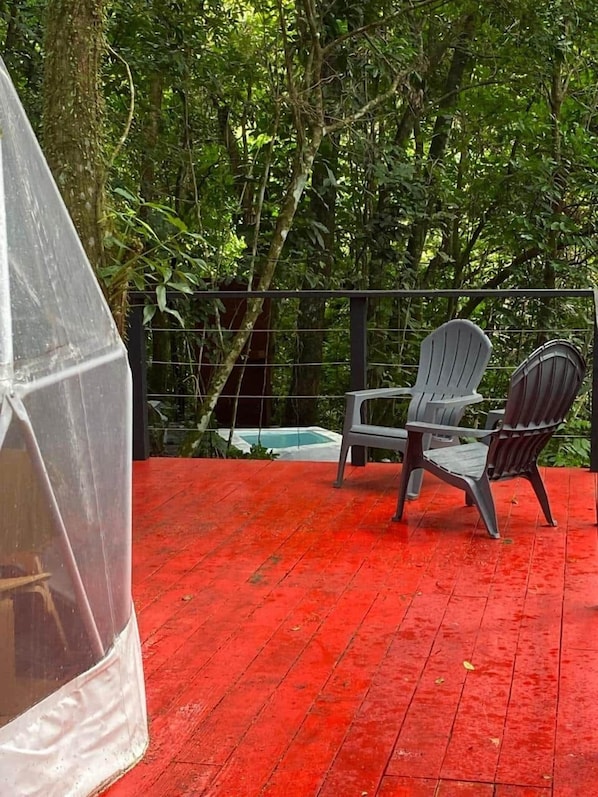 Un espacio exterior techado con piso rojo, dos sillas y una piscina al fondo.