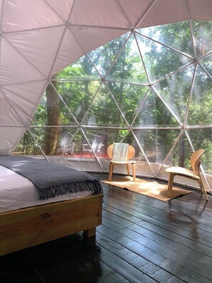 Una carpa con una cama de madera y dos sillas, rodeada de árboles.