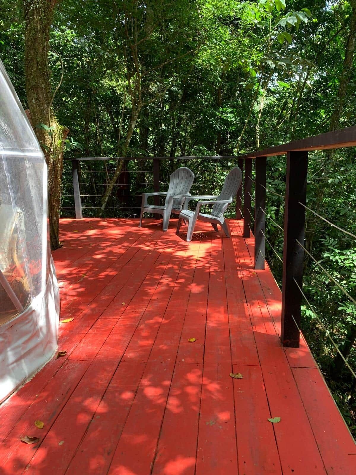 Una terraza de madera roja con dos sillas Adirondack, rodeada de follaje verde exuberante.