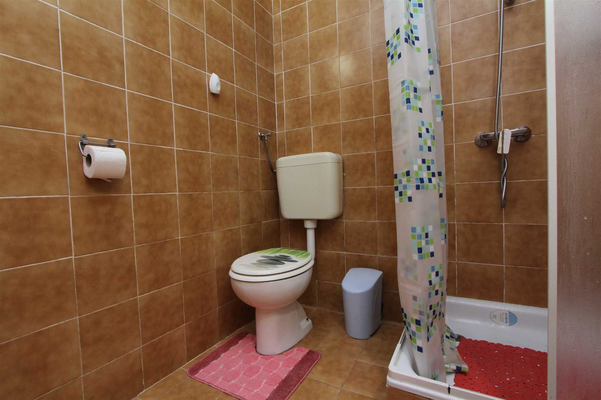 A bathroom with brown tiles, a toilet, a shower with a curtain, and a trash bin.