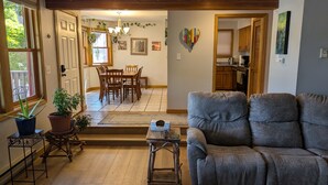 A living room with a grey sofa, wooden dining table, and chairs. There is a small wooden table with a tray and a potted plant. The room has wooden flooring and a large window with blinds. - In-town Durango Getaway | Pool | Hot Tub (Durango)
