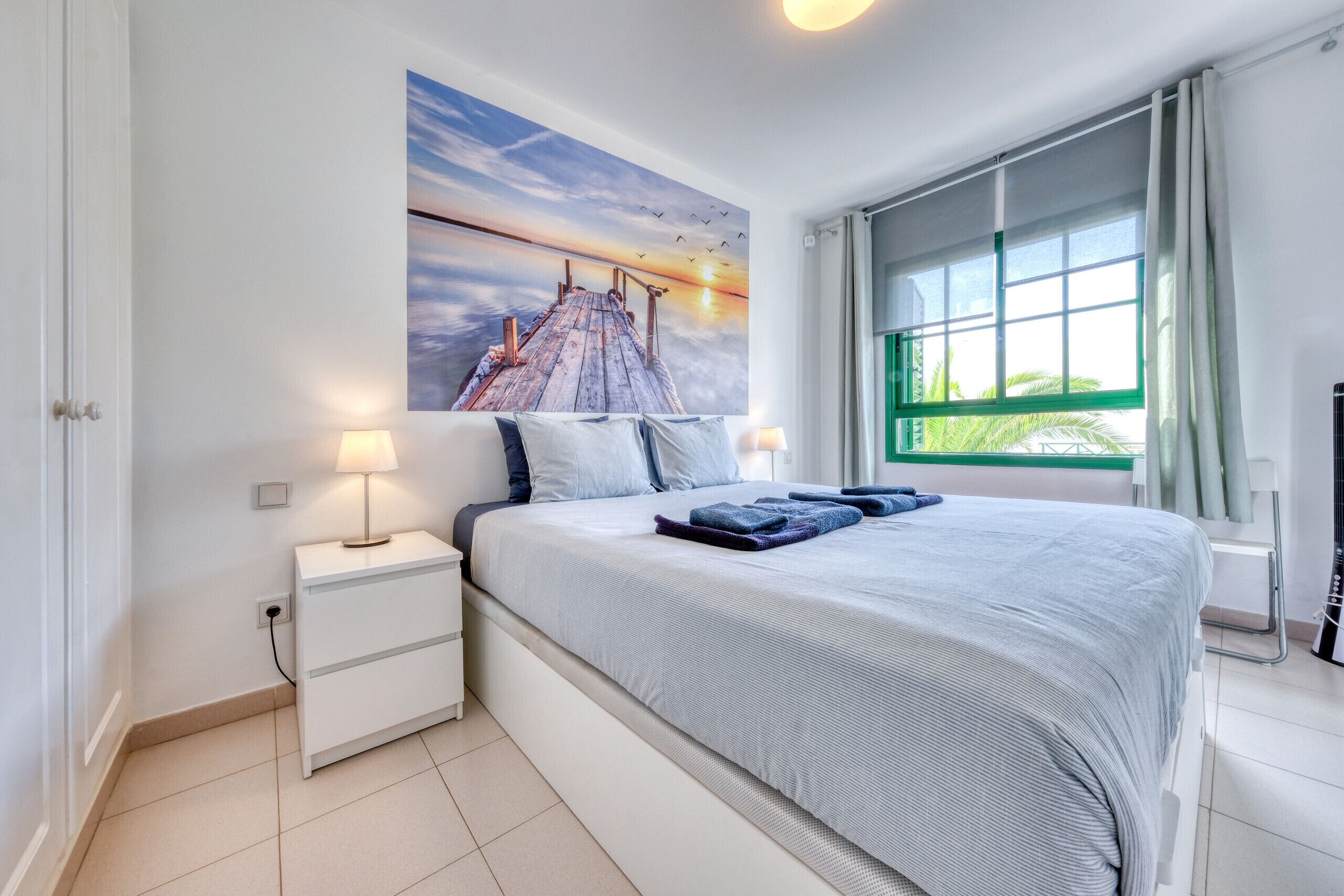 A bedroom with a large bed, a nightstand, a wall-mounted lamp, and a window with curtains.