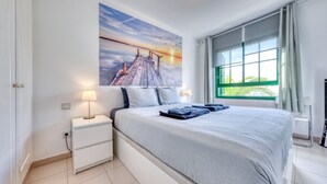 Un dormitorio con una cama grande, una mesita de noche, una lámpara de pared y una ventana con cortinas.