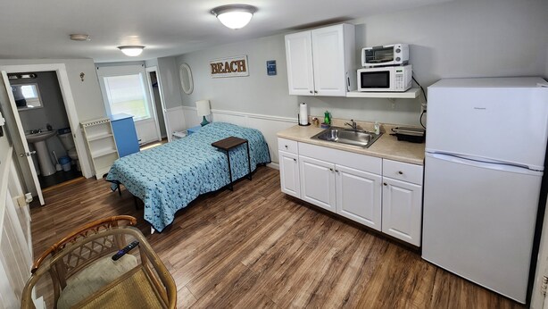 A compact kitchen with a microwave, sink, and refrigerator. A bed with a blue floral bedspread is visible in the background.