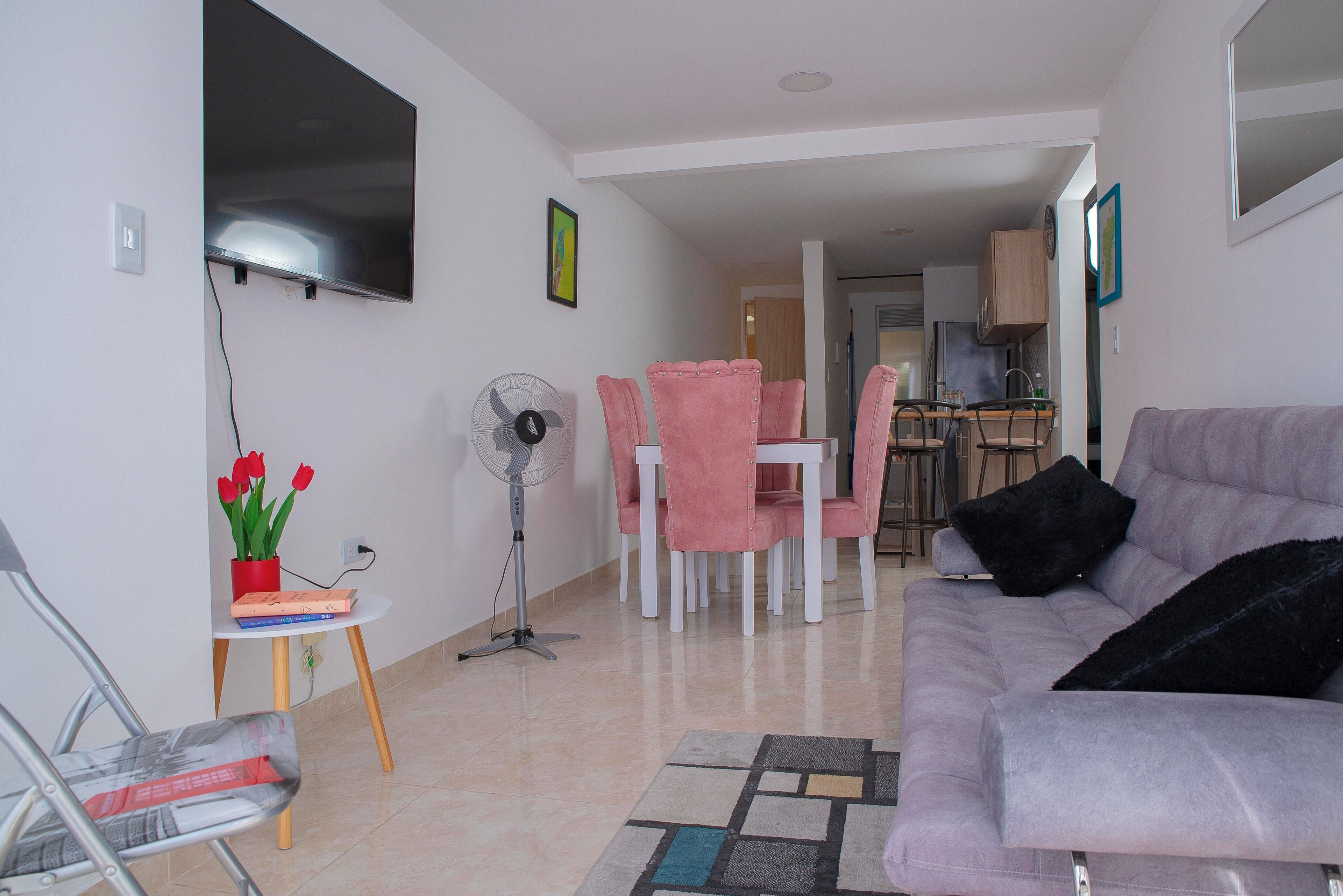 Un salon comprenant un canapé, une table à manger avec des chaises, un téléviseur fixé au mur, un ventilateur et une petite table sur laquelle est posée une plante.