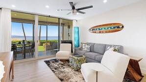 A modern living room with a sofa, armchairs, and a coffee table. There is a large window with a view of the beach.