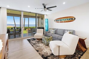 A modern living room with a sofa, armchairs, and a coffee table. There is a large window with a view of the beach.