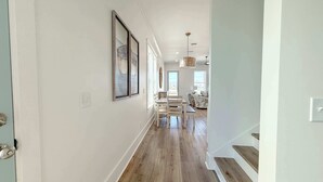 A hallway with wooden flooring, white walls, and framed artwork. There is a dining area with a table and chairs in the background.