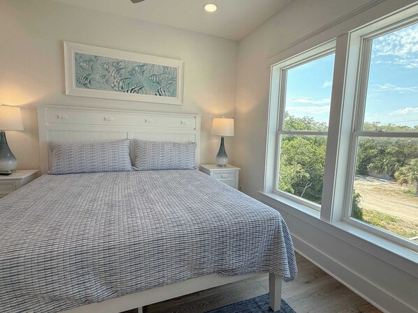 Un dormitorio con una cama grande, una mesita de noche con lámpara, una obra de arte enmarcada sobre la cama y una ventana con vistas a zonas verdes.