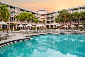 A hotel pool area with multiple lounge chairs and umbrellas, surrounded by palm trees and a multi-story building with balconies. - Pool View 2 King | Golf, Beach & Tiki Bars (Stuart)