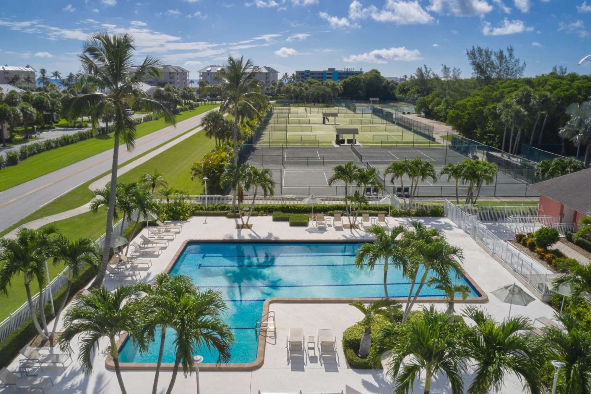 Ein Swimmingpool mit Liegestühlen und Palmen, umgeben von einer gepflegten Rasenfläche und einem Tennisplatz.