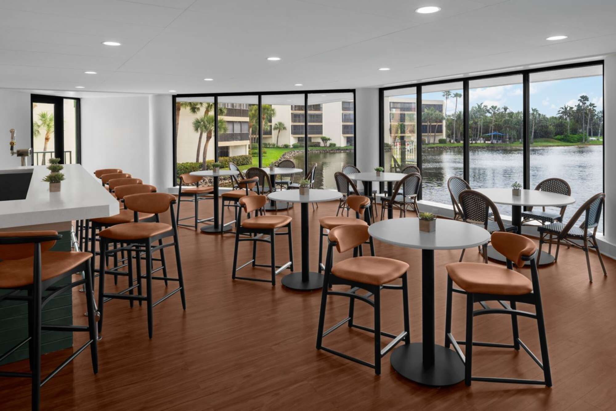A modern indoor dining area with multiple tables and chairs, large windows offering a view of a body of water and buildings.