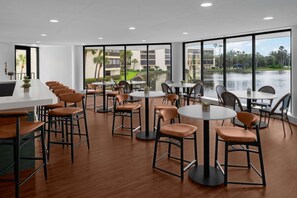 Un comedor moderno con varias mesas y sillas, grandes ventanales que ofrecen vista a un cuerpo de agua y edificios.