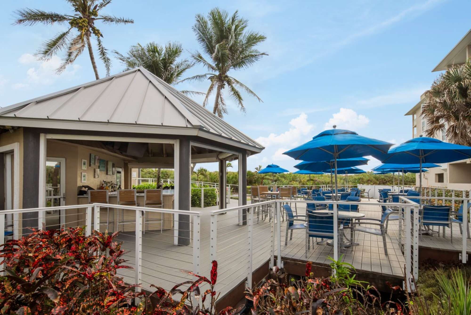 A covered outdoor seating area with tables and chairs, surrounded by palm trees and blue umbrellas.