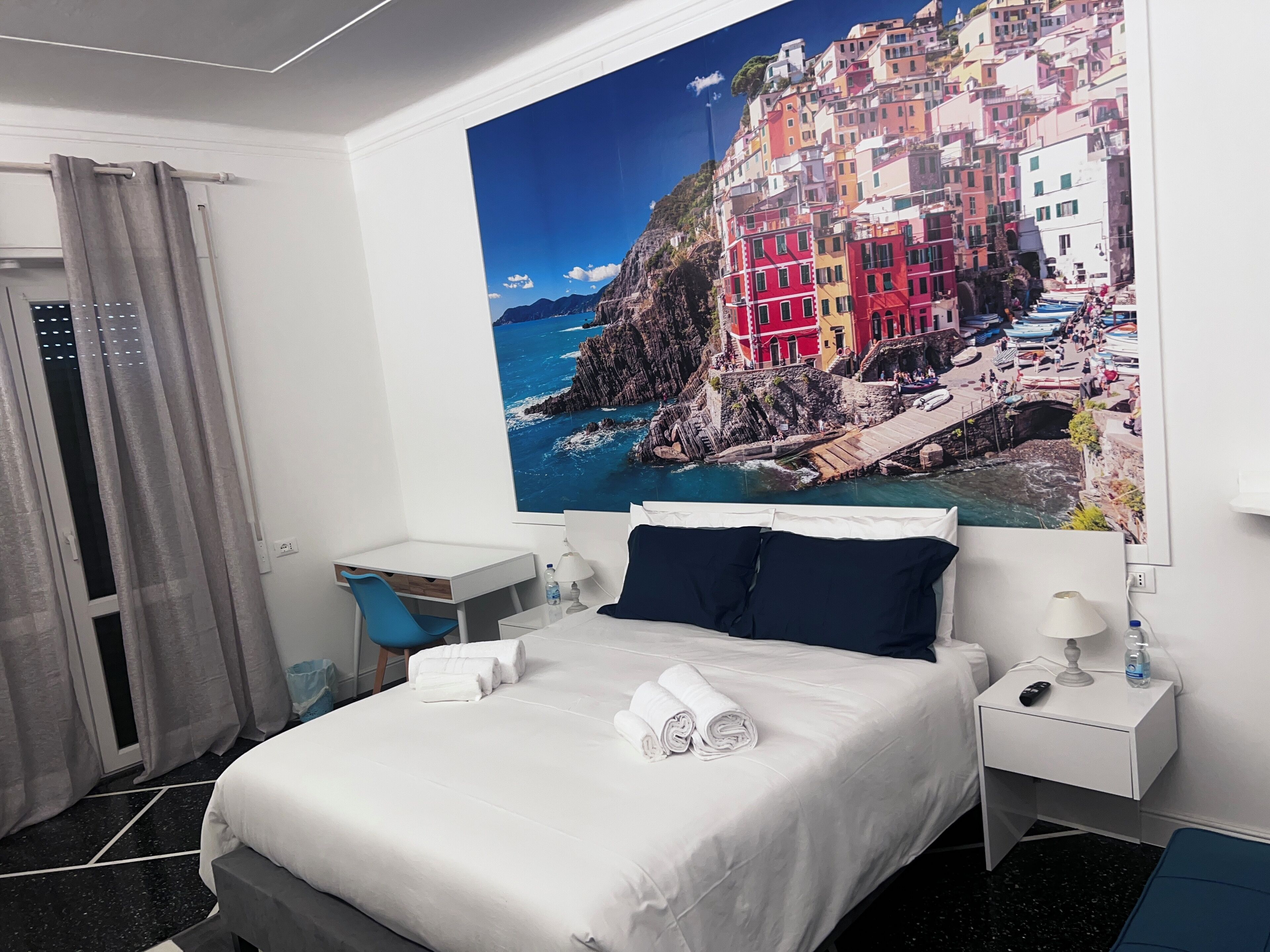 A hotel room with a bed, a desk, a chair, a nightstand, a lamp, and a large painting of a coastal town.